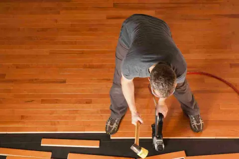 A man is installing waterproof flooring, using a nail gun and mallet to secure wooden planks.