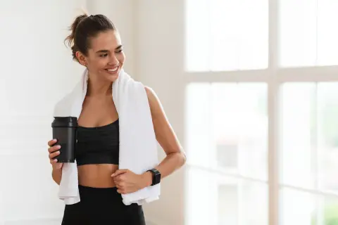 Lächelnde Frau mit Handtuch und Shaker nach dem Training. Sie fragt sich, wie viel Kreatin pro Tag für optimale Ergebnisse.