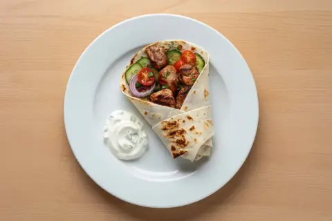 Kebab w tortilli z warzywami i sosem na białym talerzu.