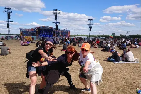 Trzy kobiety pozują na tle sceny festiwalowej Pol'and'Rock. Tłum ludzi siedzi na trawie, ciesząc się pogodą i oczekując na występ.