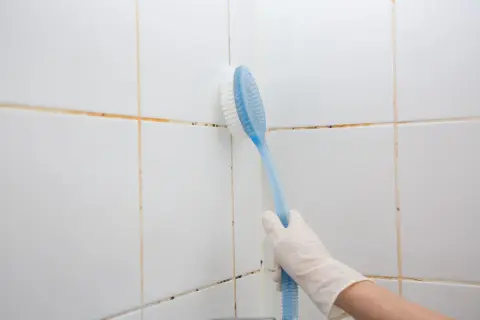 Mano con guanto bianco che pulisce le fughe della doccia con una spazzola blu.