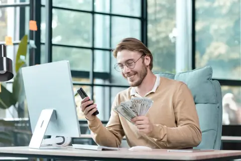 Uśmiechnięty mężczyzna z okularami trzyma telefon i pieniądze. Zastanawia się, co wpisać w tytule przelewu dla kolegi.