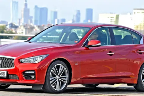 Czerwony infiniti samochód z kierowcą za kierownicą, na tle miejskiej panoramy.