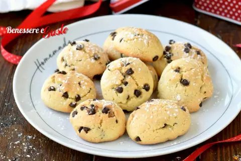 Biscotti con gocce di cioccolato spolverati di zucchero a velo su un piatto bianco, pronti per essere gustati.