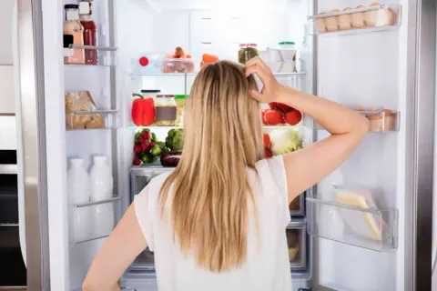 Donna confusa davanti al frigo pieno di cibo, con il desiderio di mangiare e non ingrassare, ma lo stress la blocca.