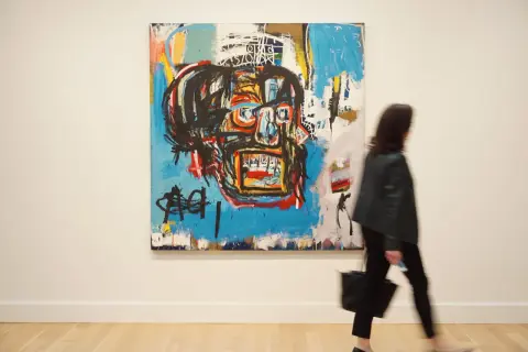 A woman walks past a vibrant, abstract painting, a striking example of Basquiat's famous paintings, featuring a bold, stylized face.