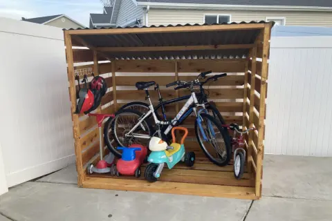 A DIY bike rack made of wood stores two adult bikes, a tricycle, and two ride-on toys. Helmets hang on a hook.