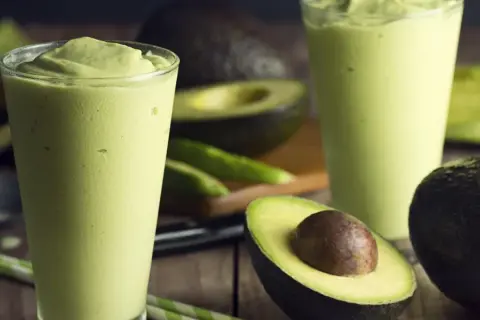 Dwa szklanki kremowego smoothie z awokado, idealna przekąska na upalne dni. Obok leżą świeże awokado i zielone słomki.