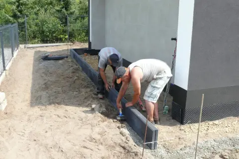 Dwóch mężczyzn układa obrzeża pod kostkę. To kluczowy etap, jak zrobić suchy beton pod kostkę, by nawierzchnia była trwała.