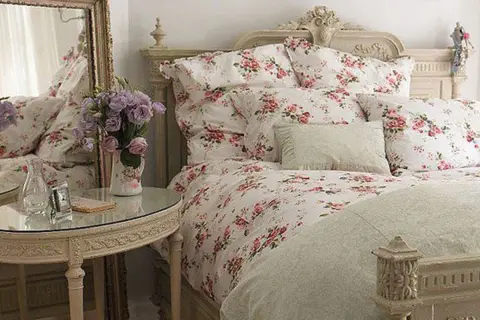 En säng med blommig shabby chic inredning, prydd med rosor och lavendel. Ett runt spegelbord med en vas full av blommor.