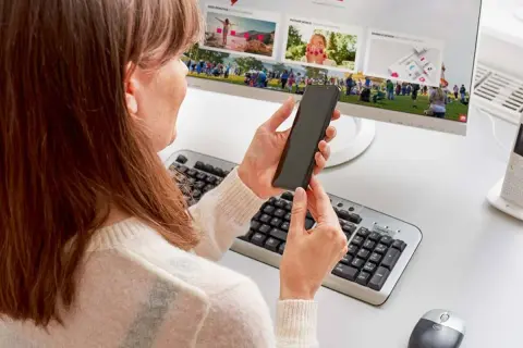 Kobieta trzyma telefon, szukając informacji, jak podłączyć telefon do monitora. Na ekranie komputera widać zdjęcia.
