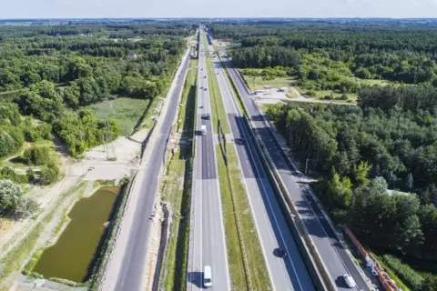 Szeroka autostrada S7 Warszawa-Grójec, otoczona lasami, z kilkoma samochodami w ruchu.