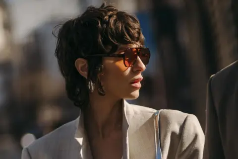 Frau mit einem frechen Pixie Cut, Sonnenbrille und Blazer.