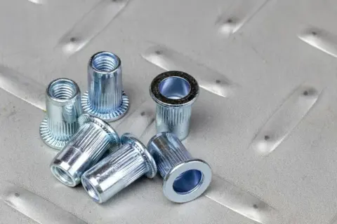 Kilka metalowych nitonakrętek leży na chropowatej, metalowej powierzchni.