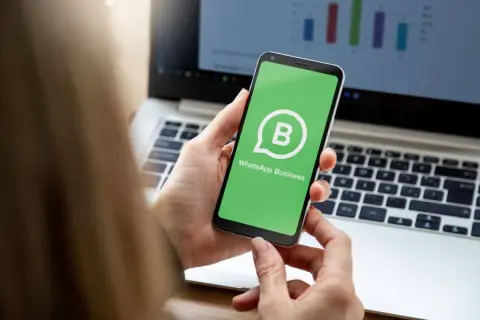 Dłoń trzyma smartfon z zielonym ekranem i logo WhatsApp Business. W tle widać laptop z wykresem.