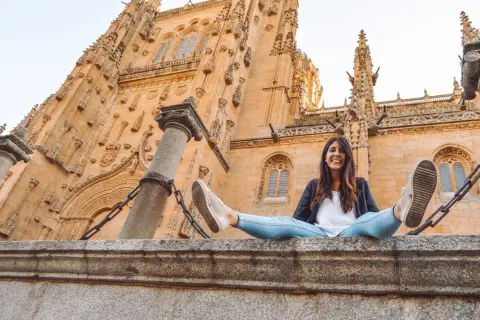 Una mujer sonríe sentada en un muro de piedra frente a la imponente fachada de la Catedral de Salamanca, un lugar clave que ver en Salamanca.