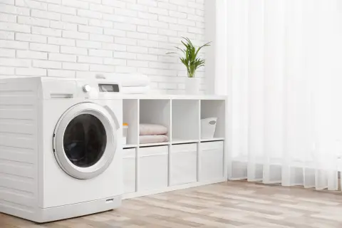 Weiße Waschmaschine mit beladener Trommel steht neben einem Regal mit Handtüchern und Aufbewahrungsboxen. Die Waschmaschine wandert nicht, sie steht fest.