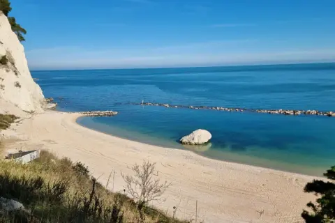 Spiaggia dorata e mare cristallino sulla Riviera del Conero, con una scogliera bianca a picco sull'acqua e un grande masso solitario.