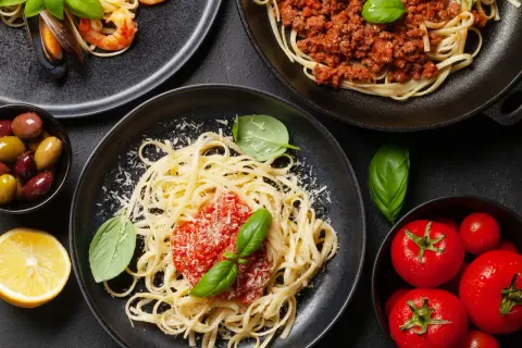 Pyszne spaghetti z sosem pomidorowym, posypane startym serem, obok krewetki z małżami, mięso mielone i świeże pomidory.