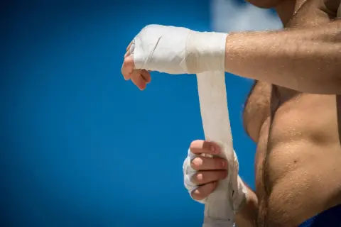 A boxer wraps his hands, preparing for a fight under a bright blue sky.