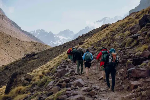 Grupa turystów wspina się kamienistym szlakiem na zboczu gór w Maroku. W tle widać ośnieżone szczyty, prawdopodobnie Jebel Toubkal.