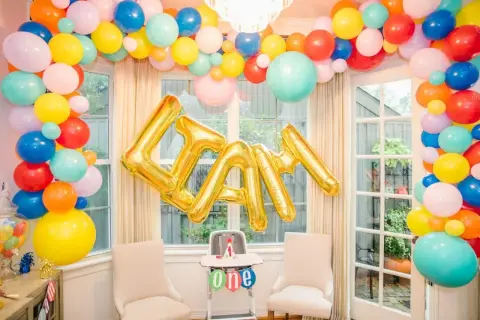 Złote balony na roczek tworzą napis "LIAM" na tle kolorowej girlandy z balonów.
