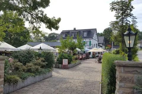Gemütlicher Biergarten mit Sonnenschirmen und Gästen. Ein Schild weist auf den "Kleine Schweiz Velbert Rundweg" hin.