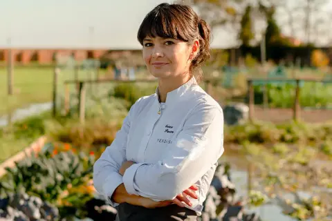 Chiara Pavan, chef con grembiule e giacca bianca, sorride con le braccia conserte in un orto.