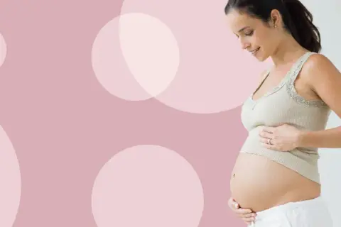 Uśmiechnięta kobieta w ciąży dotyka brzucha. Waga dziecka w 27 tygodniu ciąży jest już znacząca.