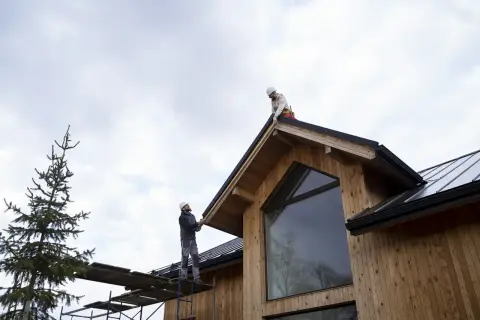 Dwóch budowlańców na dachu drewnianego domu, pracujących przy okapie. W tle sosna i niebo. Drewno c27.