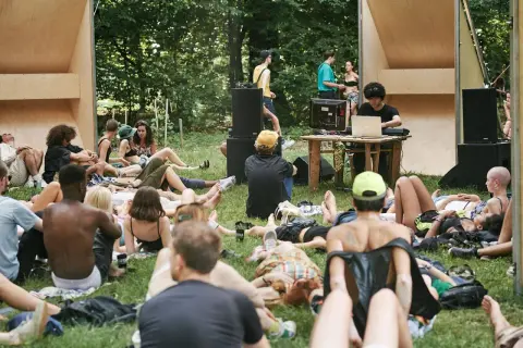 Un gruppo di persone rilassate sull'erba, ascoltando musica a un butik festival. Un DJ mixa con un laptop, circondato da altoparlanti e una struttura in legno.