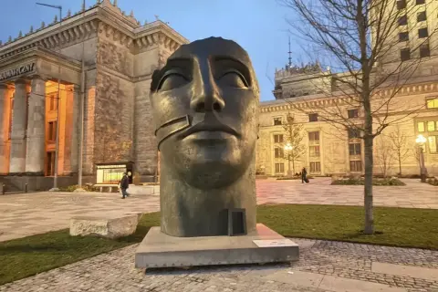 Głowa rzeźby Igora Mitoraja "Luci di Nara" na tle Pałacu Kultury i Nauki w Warszawie.