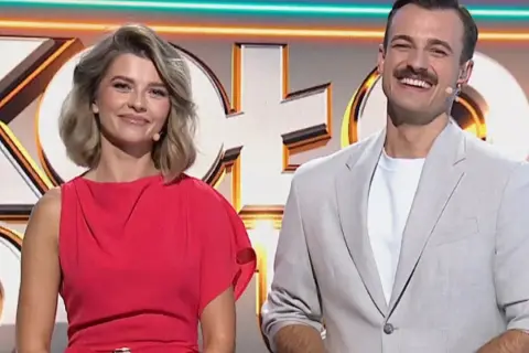 Uśmiechnięci prowadzący Koło Fortuny: ona w czerwonej sukience, on w jasnej marynarce.