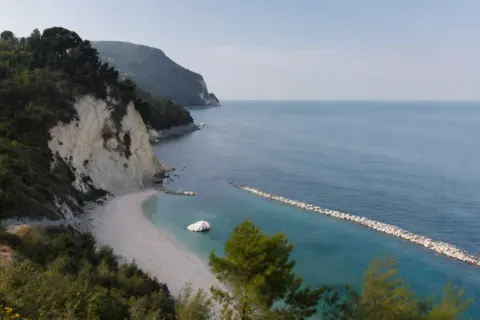 Scogliera verdeggiante e spiaggia bianca a Numana, con il lungomare che si estende verso il mare azzurro.