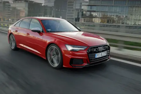 Czerwone, miękka hybryda Audi S6 sunie po drodze, za kierownicą siedzi kierowca w okularach.