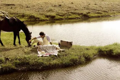 Pareja en un picnic junto a un río, con un caballo pastando. Un momento idílico para disfrutar de rutas en caballo y la naturaleza.