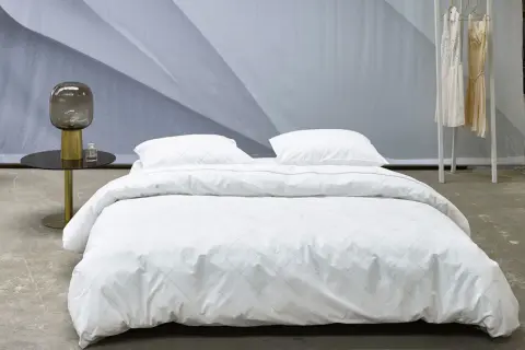 Ein Bett mit weißer Perkal-Bettwäsche, gesteppt mit feinen Linien. Daneben ein Nachttisch mit Lampe und Parfüm.