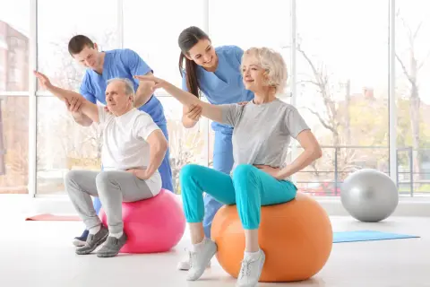 Rehabilitacja dla seniorów: starsza pani i pan ćwiczą z fizjoterapeutami na piłkach gimnastycznych, poprawiając kondycję.