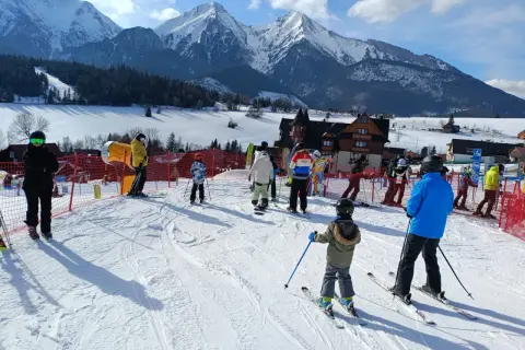 Ludzie na nartach i snowboardach w słoneczny dzień w ski centrum Strachan, z majestatycznymi ośnieżonymi górami w tle.