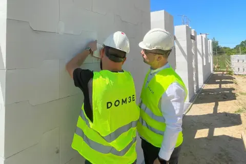 Dwóch mężczyzn w kamizelkach odblaskowych na placu budowy sprawdza postęp prac przy ścianie z białych pustaków.