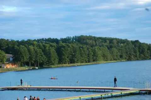 Spokojne jezioro Sierakowo z drewnianymi pomostami, na których stoją ludzie. W oddali widać kajakarzy i zalesiony brzeg.