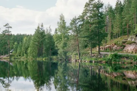 Spegelblank sjö med skog och en liten stuga vid stranden, kanske en del av Malingsbo herrgård.
