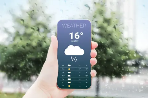 Dłoń trzyma smartfon z aplikacją pogodową pokazującą deszcz i 16 stopni. Za oknem widać krople deszczu.