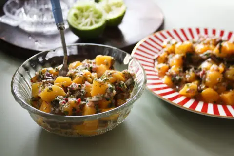 En skål med färgglad mangosalsa, med lime och en röd-vit randig tallrik i bakgrunden.