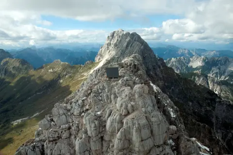 Bivacco Luca Vuerich, un cubo scuro su una cresta rocciosa, domina un panorama alpino mozzafiato sotto un cielo nuvoloso.