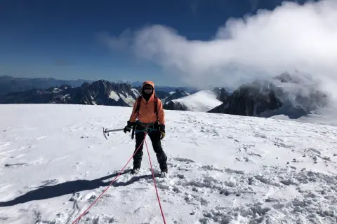Une alpiniste, équipée pour l'ascension d'une montagne, se tient sur un sommet enneigé, le regard tourné vers l'horizon.