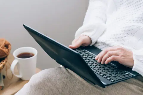 Osoba w białym swetrze pisze na laptopie, obok kubek z kawą i koszyk z włóczką.