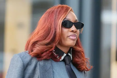 Frau mit feuriger, **cooler Haarfarbe** und stylischer Sonnenbrille trägt einen grauen Blazer mit Krawatte und glitzerndem Kragen.