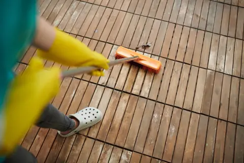 Persona con guantes amarillos y chanclas blancas usa cepillo para quitar manchas de vinagre en el suelo de madera.