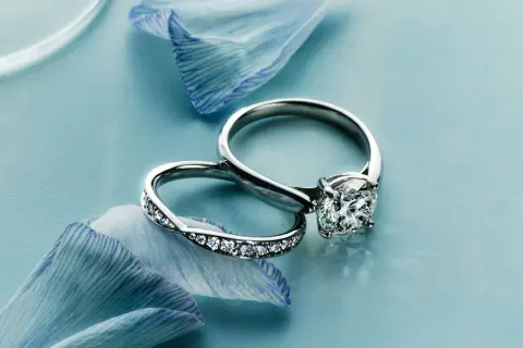 Dos anillos de compromiso, uno con un gran diamante y otro con pequeñas gemas, descansan sobre pétalos de flor azul. Un anillo casado para siempre.